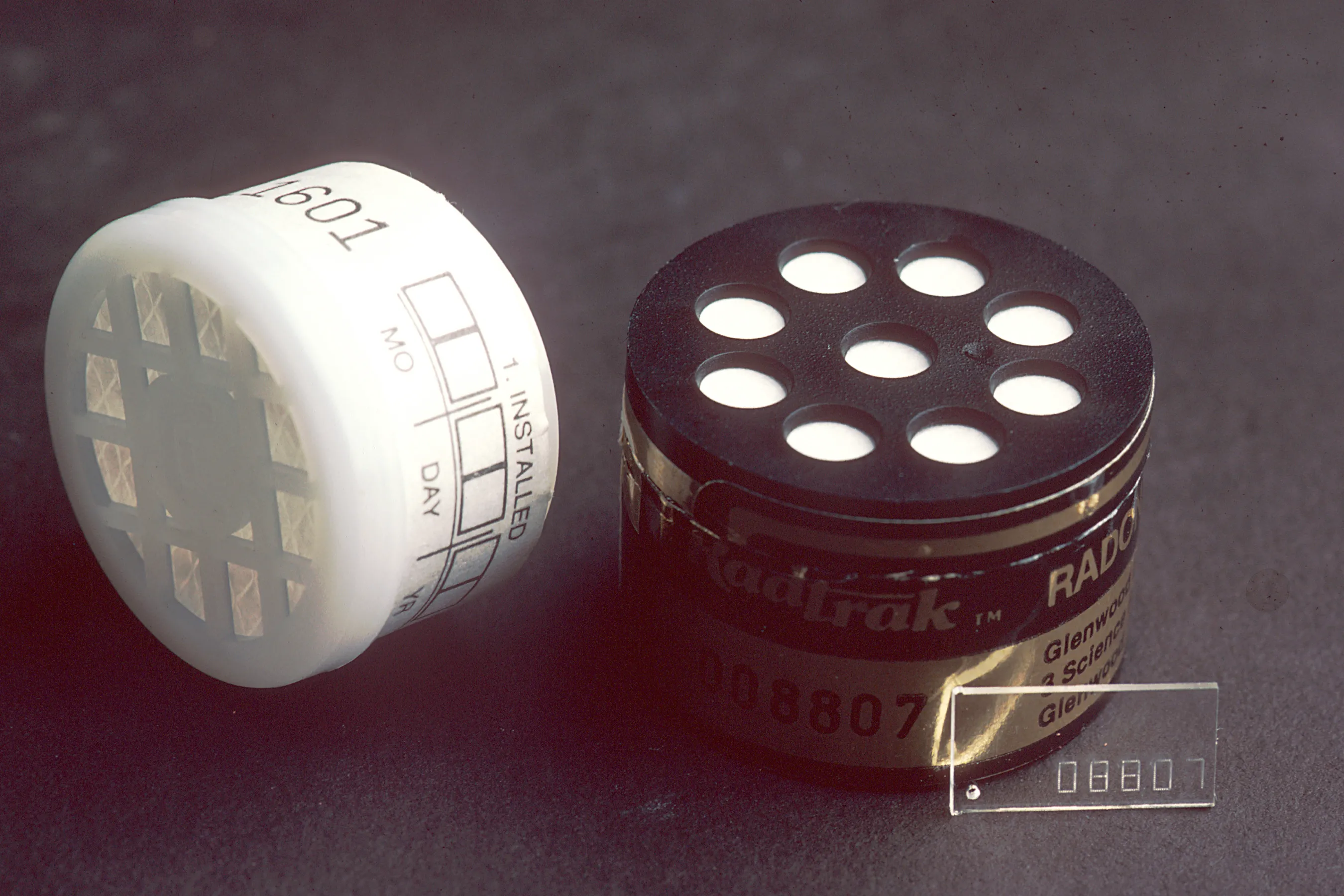 Two radon test devices and a small numbered tag on a dark surface